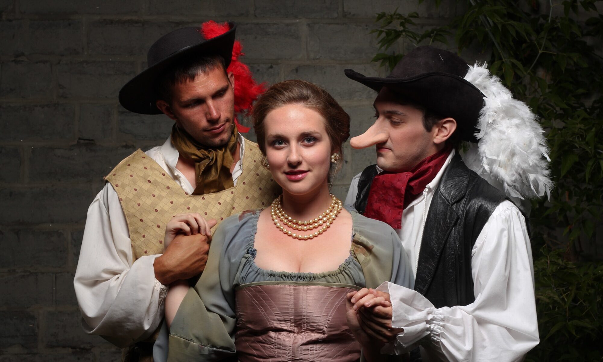 Two men in feather-adorned hats and period costumes look fondly at a woman in pale green and pink.